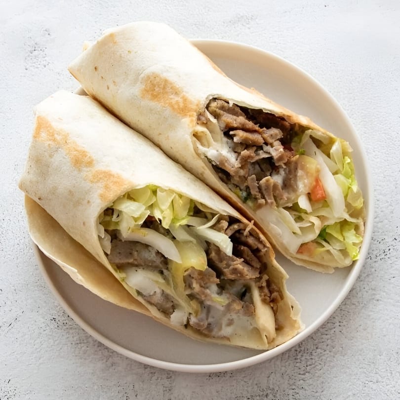 Two chicken shawarma wraps, filled with meat, lettuce, onions, and sauce, on a white plate.